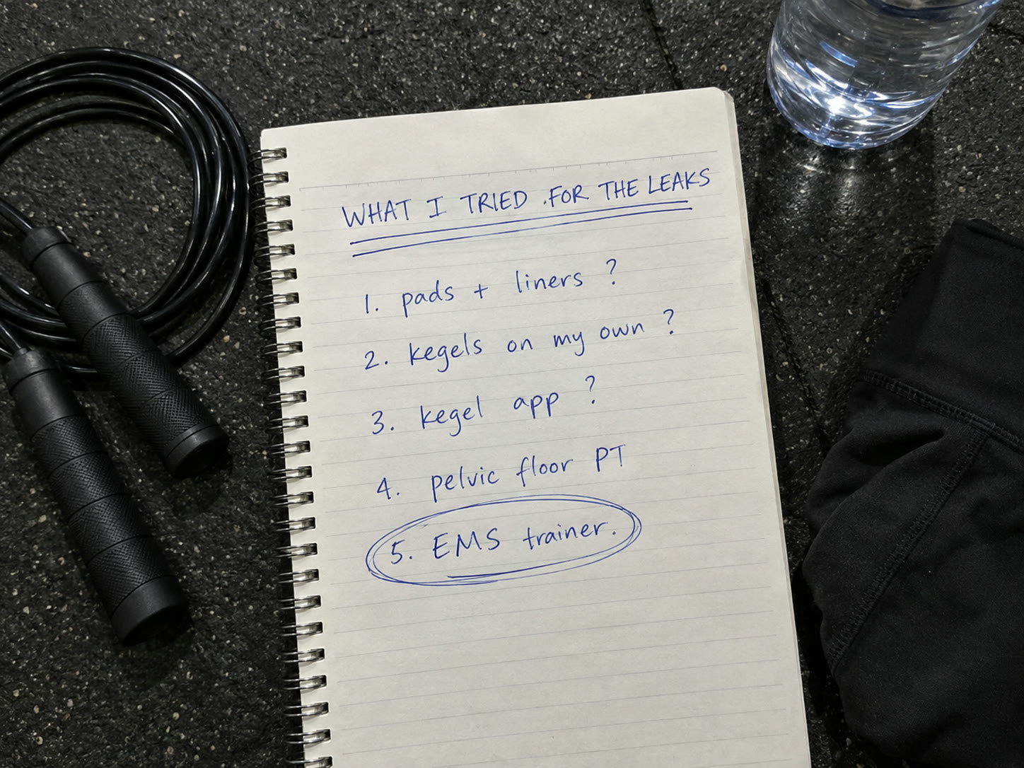 A handwritten list in a notebook on a gym floor: pads and liners, Kegels on my own, Kegel app, pelvic floor PT, EMS trainer circled