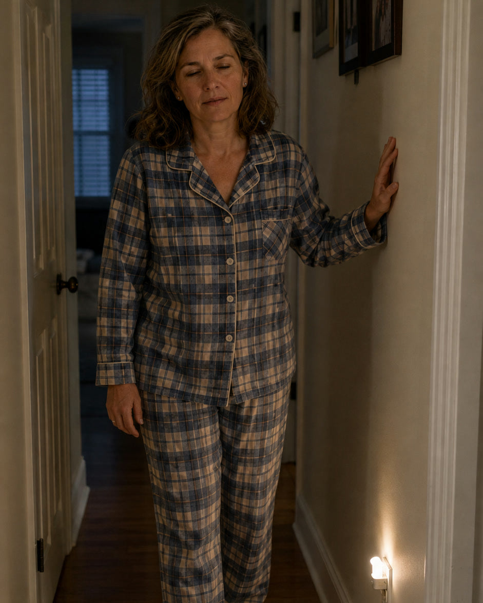 A woman in plaid pajamas walking down a dark hallway at night, one hand on the wall, a night light glowing