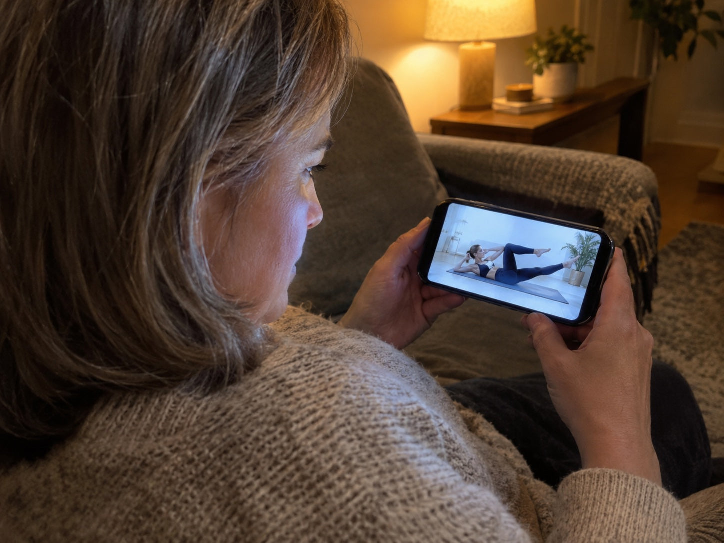 Over the shoulder: a woman on her sofa frowning at an exercise video on her phone
